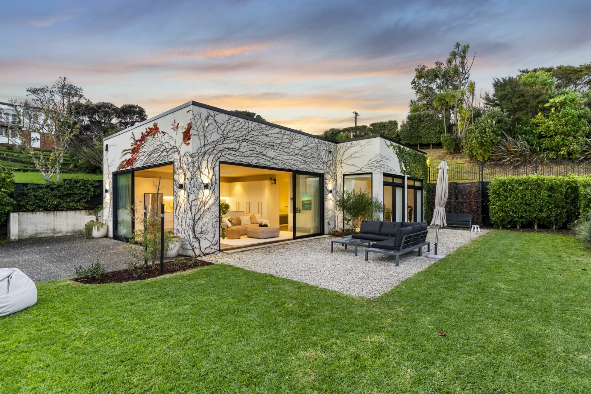 Modern Auckland home at sunset with manicured lawn, tidy hedges and styled outdoor lounge — open home ready
